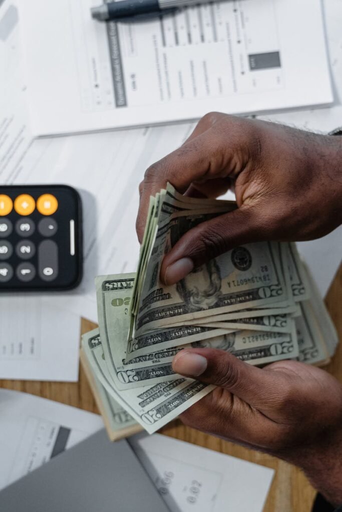 Person counting dollar bills over documents with a smartphone calculator on the desk.